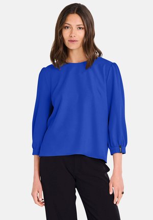 Helle blaue Bluse mit rundem Ausschnitt, drei Viertel Puffärmeln und lockerer Passform. Glattes Material mit dezenter Glanz, kombiniert mit schwarzen Hosen.