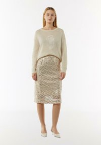 Beiger Strickpullover mit dezentem Logo, kombiniert mit einem glitzernden Bleistiftrock in Gold und Silber sowie Absatzschuhen. Glatte Textur, figurbetonte Silhouette.