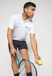 Weißes Polohemd mit dezentem Streifenmuster, Logo auf der Brust, kombiniert mit dunklen Shorts. Hält einen Tennisschläger und einen grünen Tennisball.