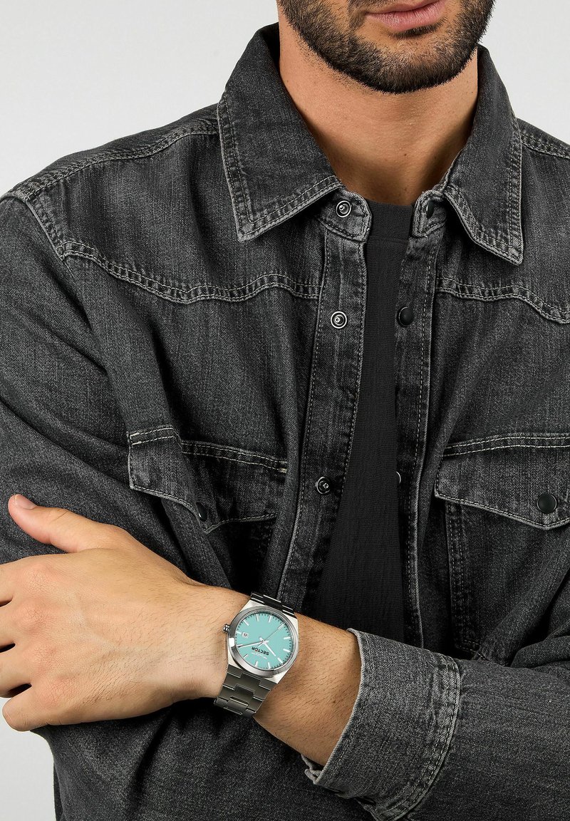 Orologio in metallo argento con quadrante turchese, cinturino a maglie e tre lancette. Indossato su un polso, parzialmente coperto da una giacca di denim scuro.