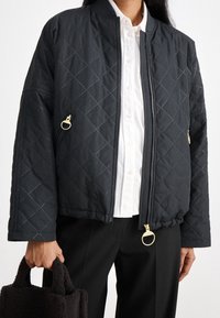 Zwarte gewatteerde bomberjack met diamantstiksel, voorzien van gouden accessoires, ritssluitingen en een geribde kraag. Gedragen over een wit overhemd.