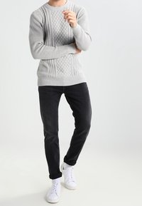 Person som bär en ljusgrå kabelstickad tröja, svarta uppvikta byxor och vita sneakers, stående med ett arm korsat och en hand nära ansiktet.