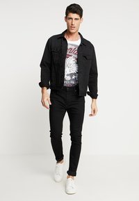 Chaqueta de mezclilla negra sobre una camiseta gráfica con un diseño de águila, combinada con jeans ajustados negros y zapatillas blancas. Atuendo simple y casual.