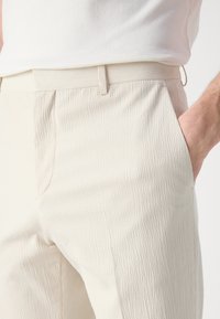 Lichtbeige, getextureerde broek van zacht materiaal, met een vlakke voorkant, zijzakken en een getailleerde pasvorm, die op de taille rust met een smalle tailleband.