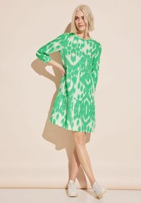 Grön mönstrad klänning med långa ärmar och utsvängd kjol. Har en abstrakt design och bärs med vita sneakers.
