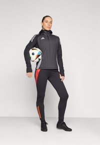 Veste de sport noire avec des accents gris, manches longues et col zippé ; pantalon noir ajusté avec un panneau latéral rouge et chaussures de sport noires.