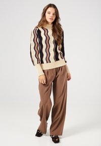Suéter de punto multicolor con patrones de olas en crema, marrón y azul marino; cuello y puños acanalados en beige. Combinado con pantalones de pierna ancha marrones.