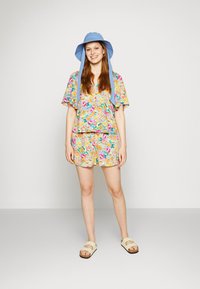 Conjunto floral colorido em duas peças, com uma blusa de manga curta e calções. Acompanha um chapéu de balde azul-claro e sandálias bege.