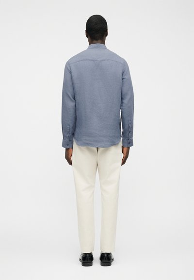 Άνδρας που στέκεται με την πλάτη προς την κάμερα φορώντας μπλε μακρυμάνικο πουκάμισο, κρεμ παντελόνι και μαύρα επίσημα παπούτσια μπροστά σε απλό φόντο.