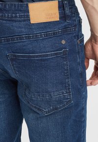 Temno modri jeans iz denima z usnjenim pecatom na hrbtu pasu z napisom "DENIM JOURNEY," standardni žepi in gladka tekstura.