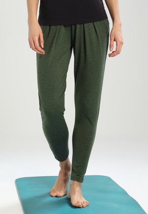 Person steht barfuß auf einer blauen Yogamatte, trägt dunkelgrüne, schmal zulaufende Hosen und ein schwarzes Hemd, die Hände entspannt an den Seiten.