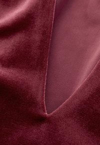 Burgundowa aksamitna tkanina o gładkiej teksturze, z głębokim wycięciem w kształcie litery V, które odkrywa kontrastową, prążkowaną podszewkę w podobnym odcieniu.