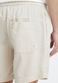 Beige teksturerte shorts med elastisk midje, med en lomme og subtilt ripet mønster. Nøytral farge på stoffet.