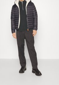 Giubbotto puffer blu navy con fodera bianca pelosa, chiusura con zip, maglione grigio sotto, pantaloni grigi e stivali neri con lacci.