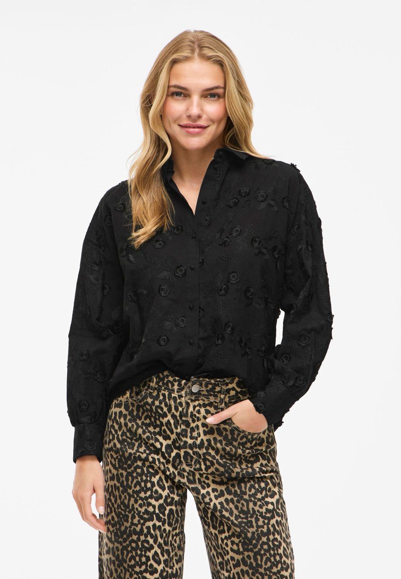 Svart broderad blus med blommönster, knäppning fram, och långa ärmar, parat med mönstrade leopardtryck byxor.