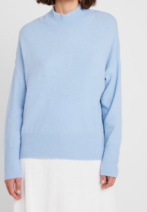 Femme portant un pull en tricot bleu clair à manches longues avec un col rond monté et des poignets et un ourlet côtelés.