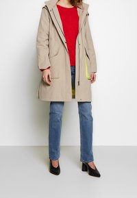 Beżowy lekkie płaszcz z kapturem, dwoma przednimi kieszeniami i żółtymi akcentami. W połączeniu z czerwoną koszulą, niebieskimi dżinsami i czarnymi butami.