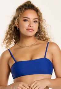 Mėlynas bandeau tipo bikini viršus su plonomis petnešėlėmis, glotnia medžiaga ir lengva blizgesiu. Pasižymi minimalistiniu dizainu su aukso tono akcentu.