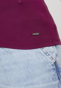Top ajustado morado hecho de una tela suave, con una pequeña etiqueta de logo plateada. Combinado con pantalones cortos de mezclilla de lavado claro con costuras visibles.