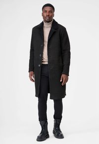 Sort frakke med imiteret pelsfor, notch-krave, knaplukning og påsyede lommer. Bæres over en beige turtleneck og sorte bukser med støvler.