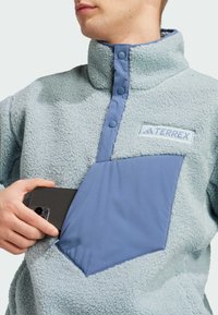 Fleece pullover i lyseblå med høj krave, der har en kontrasterende blå lomme. Inkluderer trykknaplukning og et TERREX-logo.