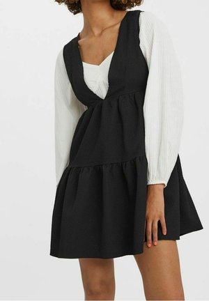 Femme portant une robe noire texturée sans manches par-dessus un chemisier blanc à manches longues, debout avec une main reposant à ses côtés.