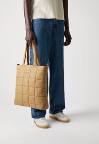 Quilted beige tote bag with padded texture and dual handles. Worn with blue denim pants and white sneakers. Minimalist design, no visible logos.
