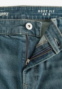 Dungaree yang berwarna biru wash sedang dengan potongan boot-cut, dilengkapi dengan kancing perak, ritsleting metal, dan detail jahitan kontras. Ukuran 34R.