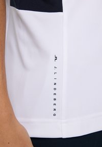 Kortärmad vit träningströja med marinblå detaljer, med en vertikal logotyp, slät textur och sömdekoration vid nederkanten.