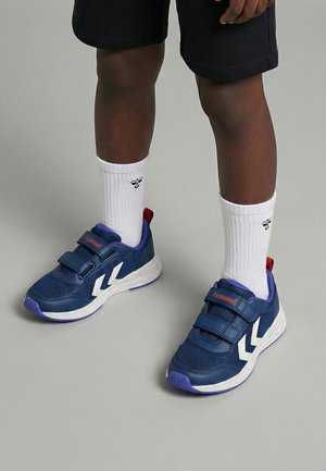 Dziecko noszące granatowe trampki z białymi paskami w kształcie litery V, białe skarpetki za kostkę z logo pszczoły oraz czarne szorty, stojące na szarej podłodze.