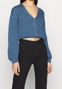 Blauwe gebreide cardigan met V-hals, cropped ontwerp, met lange mouwen en knopen. Gecombineerd met zwarte hoge taillebroek.
