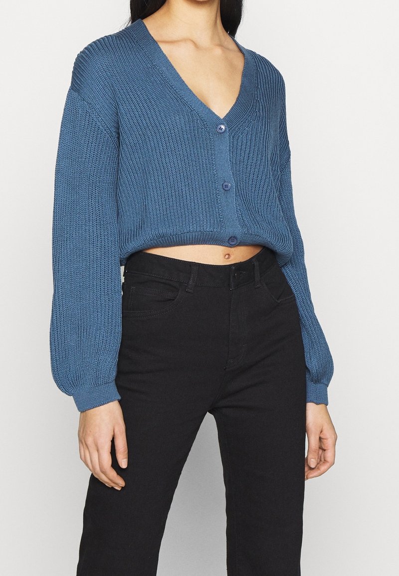 Blauwe gebreide cardigan met V-hals, cropped ontwerp, met lange mouwen en knopen. Gecombineerd met zwarte hoge taillebroek.