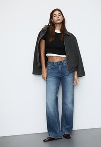 Črn kratek top z belim robom pod sivo preveliko jakno, v kombinaciji z modrimi širokimi hlačami in rjavimi čevlji. Srebrn prstan kot dodatek.