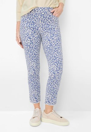 Frau trägt blaue und weiße Hose mit Leopardenmuster und beige Sneaker, steht vor einem weißen Hintergrund mit einer Hand in der Tasche.