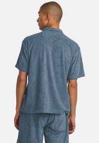 Camisa corta de manga azul con textura y un patrón floral, con cuello y un corte relajado. La tela es suave y ligeramente arrugada.