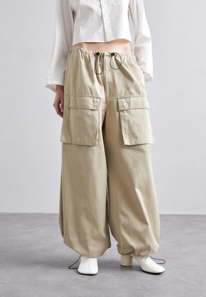 Pantalon cargo beige à jambes larges avec grandes poches avant surdimensionnées et taille à cordon, associé à des bottines blanches à talons et une chemise blanche courte.