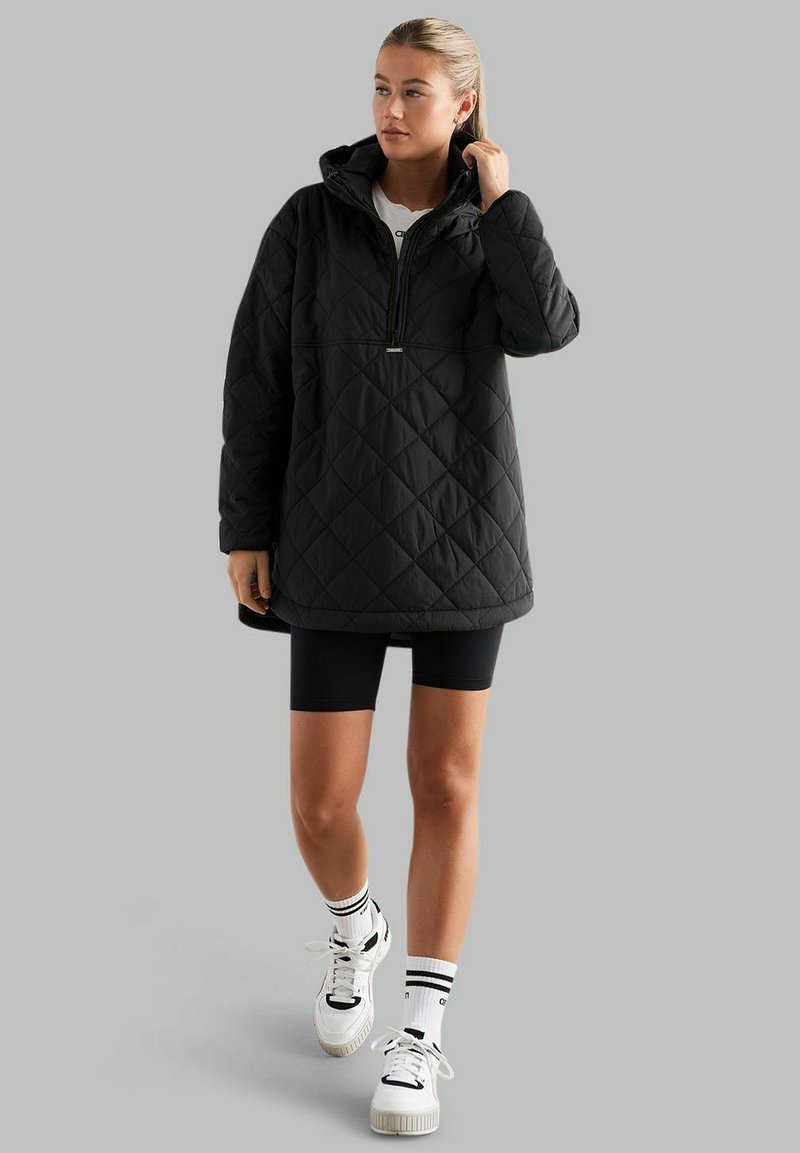 Zwart gewatteerd anorakjack met een capuchon, halve rits aan de voorkant en een kangoeroezak; gecombineerd met zwarte shorts en witte sneakers, met gestreepte sokken.