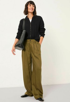 Black cable-knit cardigan with zip closure, oversized olive green trousers with pleats, and sleek black shoes. Model holds a black shoulder bag.