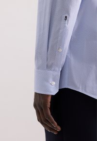 Manica di camicia a righe azzurro chiaro con piccoli motivi a diamante, bottoni bianchi e dettaglio ricamato di una rosa vicino al polsino.