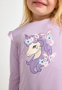 Λιλά μακρυμάνικο πουκάμισο με γραφικό μονόκερου με μωβ χαίτη, ανθισμένες λεπτομέρειες και πεταλούδες. Μαλακή υφή με βολάν στους ώμους.