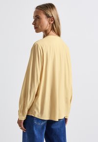 Blusa a maniche lunghe gialla chiara con un fit rilassato, texture morbida, polsini arricciati e orlo posteriore arrotondato, abbinata a jeans blu.