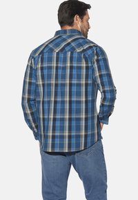 Mann in einem karierten Langarmhemd in Blau, Schwarz und Weiß und blauen Jeans, der mit dem Rücken zum Betrachter vor einem schlichten Hintergrund steht.