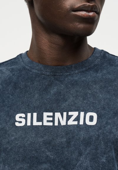 Κοντινό πλάνο ενός ατόμου που φοράει ξεθωριασμένο σκούρο μπλε πουκάμισο με λευκό κείμενο "SILENZIO" στο στήθος, μερική προβολή προσώπου και λαιμού.