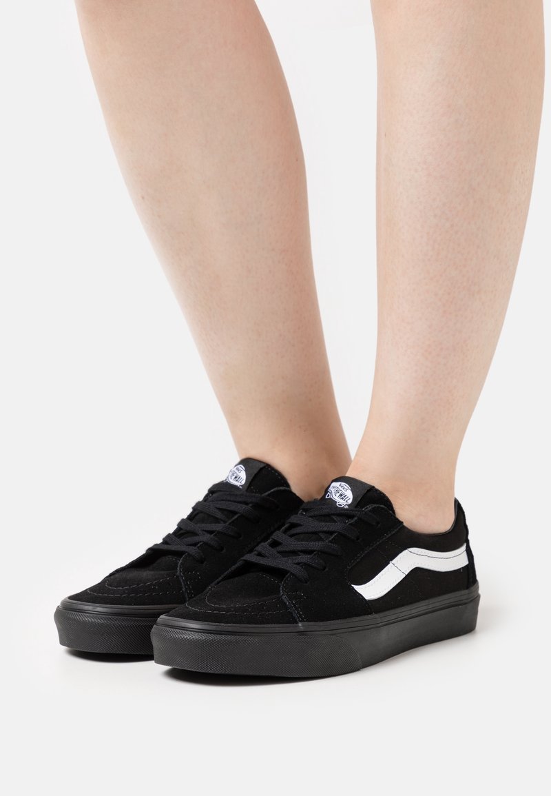 Baskets basses noires avec bande blanche sur le côté, portées pieds nus, vues de face et de profil sur fond blanc.