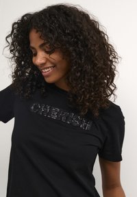 T-shirt en coton noir avec "CHERISH" en sequins noirs brillants, manches courtes, col rond et coupe décontractée. Cheveux bouclés partiellement visibles.