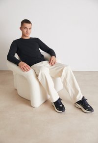 Melna garām piedurknēm krekls, krēmkrāsas platās bikses un melni sporta apavi atrodas uz teksturēta baltā krēsla minimālistiskā vidē.