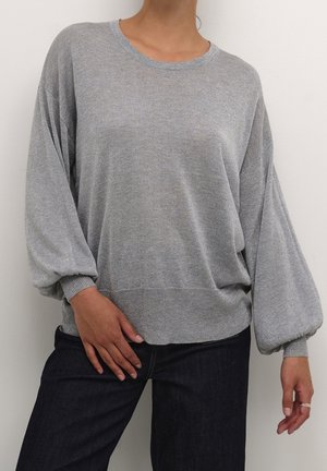 Personne portant un pull en maille gris clair avec des manches bouffantes et un jean foncé, debout devant un mur blanc uni.
