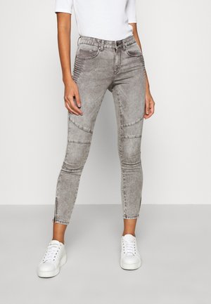 Persona vistiendo jeans ajustados estilo motorista en gris desgastado y zapatillas blancas, de pie contra un fondo claro y sencillo.