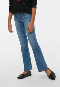 Osoba v světle modrých džínách typu bootcut, černých balerínách a černém dlouhém tričku stojí před jednobarevným bílým pozadím.