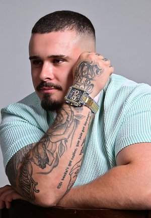 Square, gold and silver wristwatch with a black face and patterned hour markers. The person wears a green striped shirt with visible tattoos.